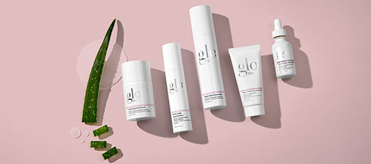 Aloe leaf with cut aloe pieces beside five white-and-pink glo skincare bottles and a dropper on pink background