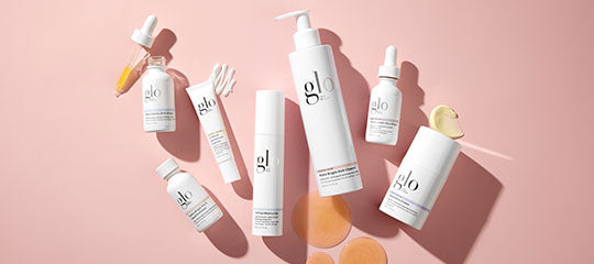 Assortment of glo skincare bottles and droppers with round cotton pads on a pink background.