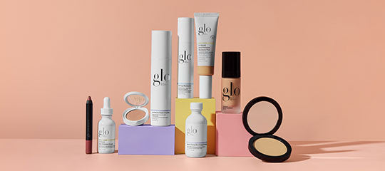 Assorted glo cosmetic bottles, tubes, compact, and dropper on pastel blocks against a peach background