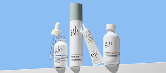 Four glo skincare products—dropper, tall pump, tube, and capped bottle—on a white ledge with blue background