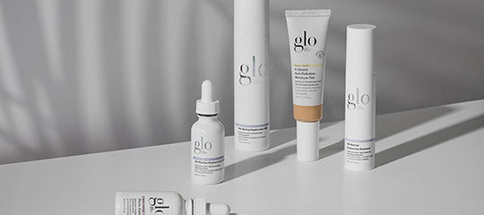 Assortment of glo skincare products—serum, tubes and bottles—arranged on a reflective white surface with soft shadows.