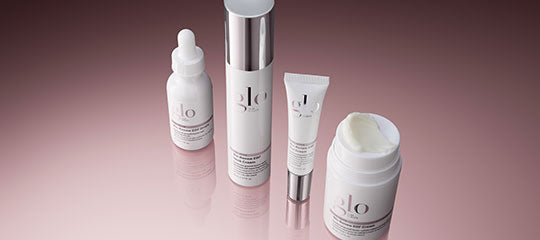 glo skincare products (dropper bottle, pump bottle, tube, open jar) on reflective pink surface