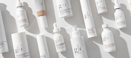 Assorted white 'glo' skincare bottles and dropper bottles laid on a pale background, casting soft shadows.