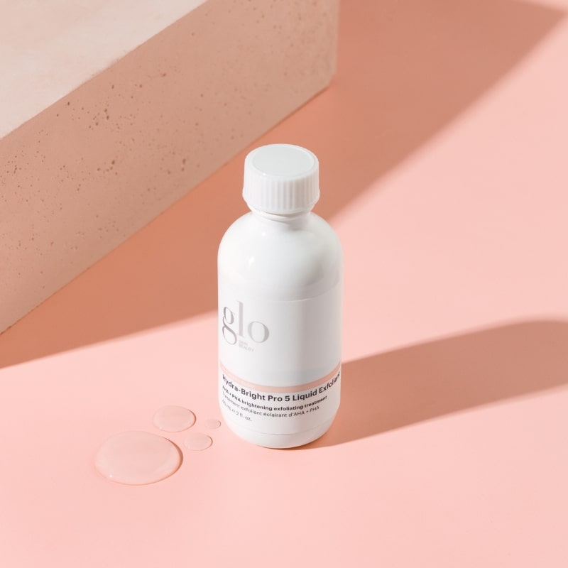 White bottle labeled glo Hydra-Bright Pro 5 Liquid Exfoliant on pink background with droplets