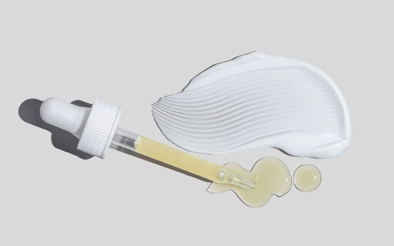 Dropper with yellow serum beside a white cream smear on a pale gray background