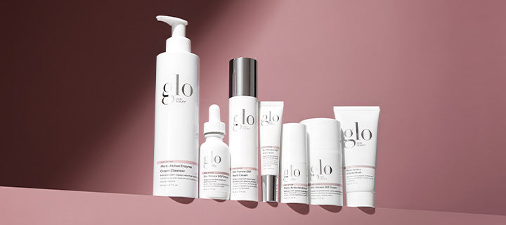 Row of white 'glo' skincare bottles, tubes and dropper on a pink backdrop, arranged on a ledge.
