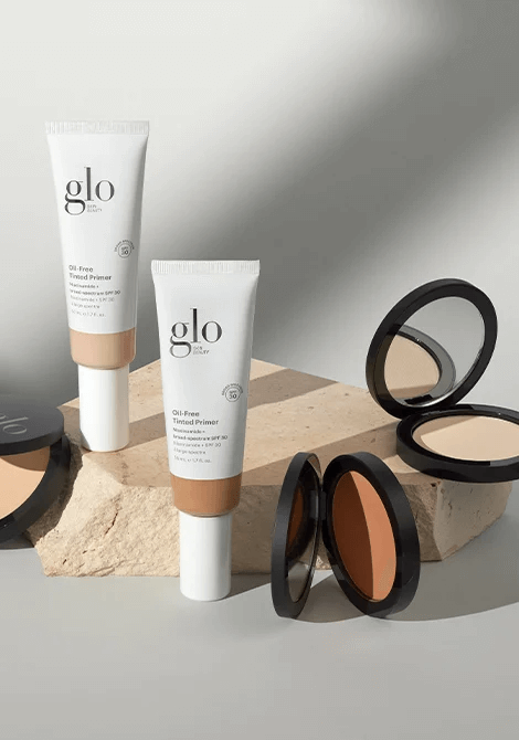 Two glo Oil-Free Tinted Primer tubes and two compact powders on a stone prop, beige shades.