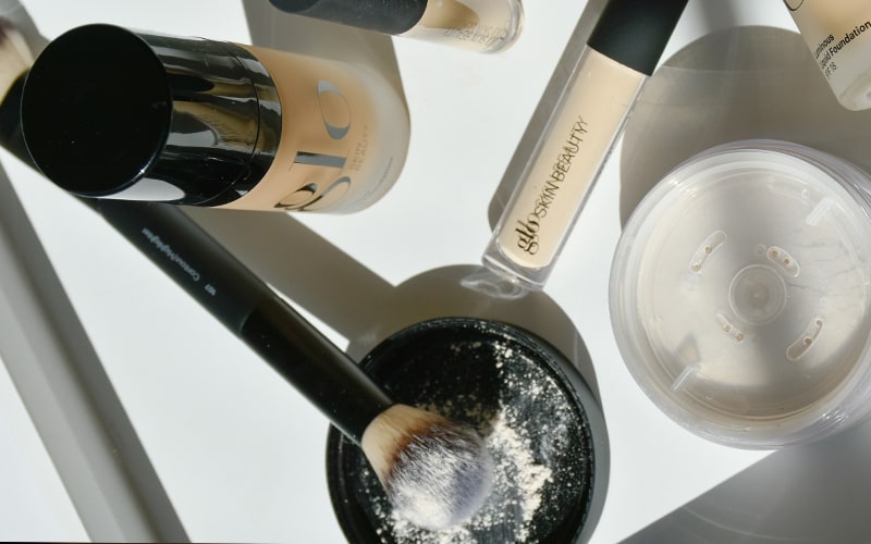 Makeup items: DIOR foundation bottle, concealer tube labeled SKINBEAUTY, loose powder and a face brush dusted with powder.