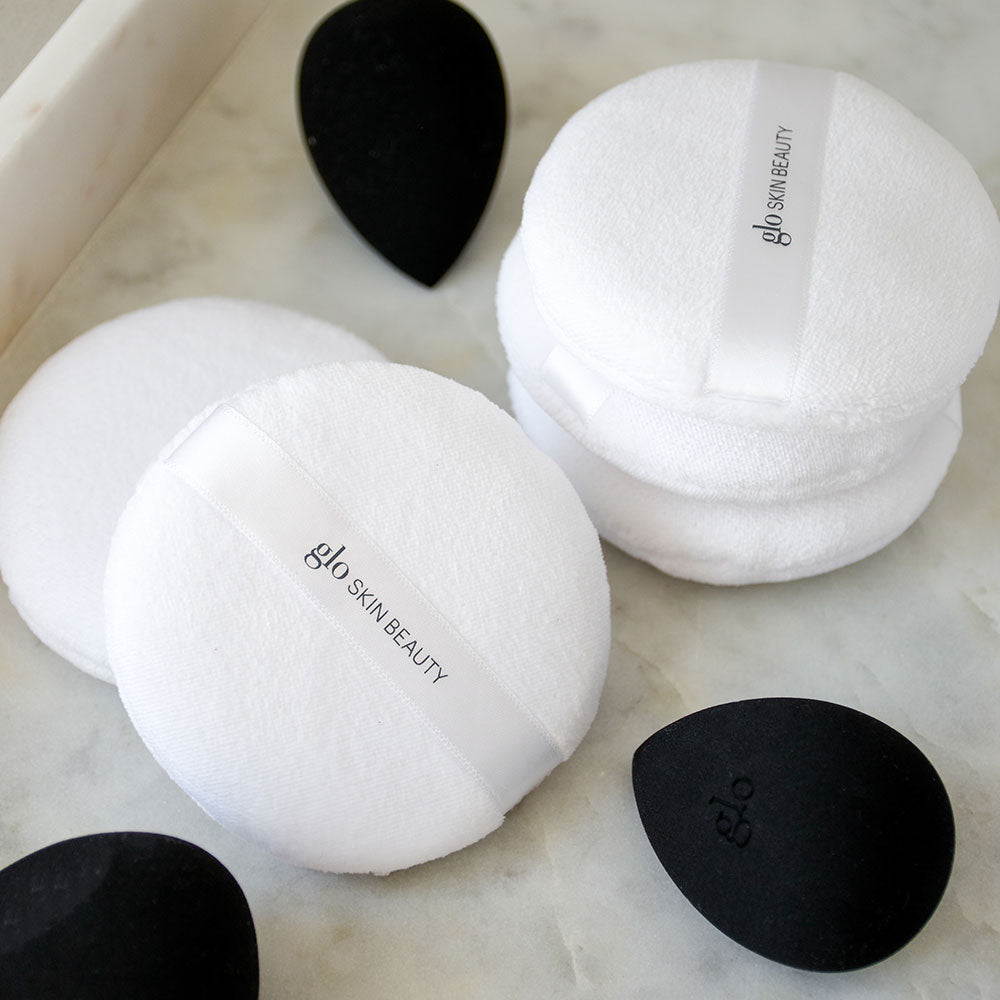 White round powder puffs with ribbon labeled glo SKIN BEAUTY and black teardrop makeup sponges embossed glo on marble surface
