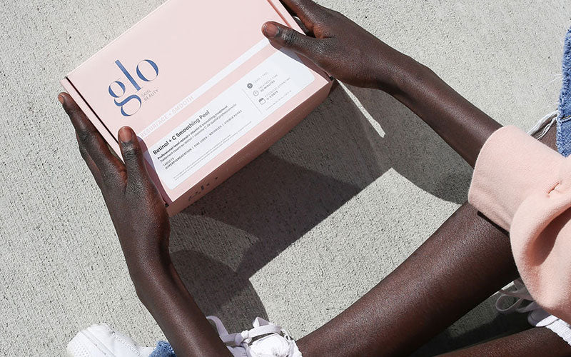 Hands holding a pink glo box labeled 'Retinol + C Smoothing Peel' on concrete background