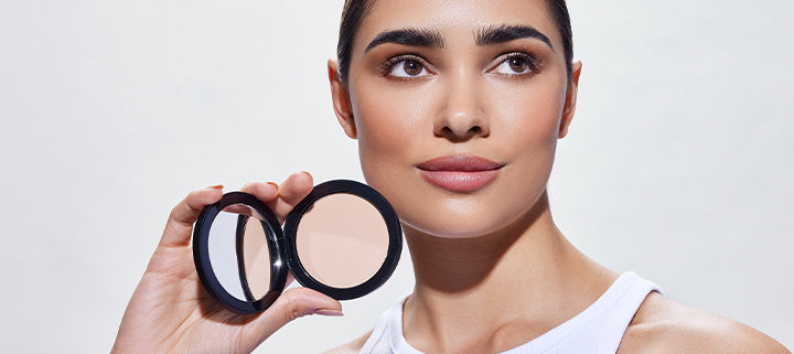 Young woman holding an open compact powder with mirror; neutral pressed powder visible, wearing a white tank top.