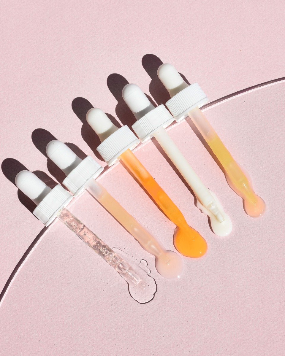 Five droppers with white caps, each dripping a distinct serum: clear, pale pink, peach, orange, and milky, on pink background