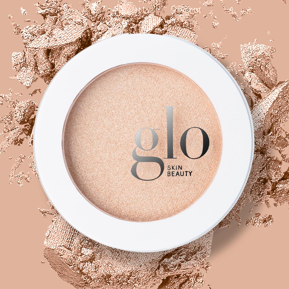 Round peach highlighter compact with glo SKIN BEAUTY logo on the lid, white rim and scattered powder background
