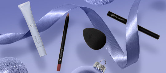 Flat lay of makeup: primer tube, lip liner pencil, black teardrop sponge, two eyeliners, lavender ribbon and ornament.