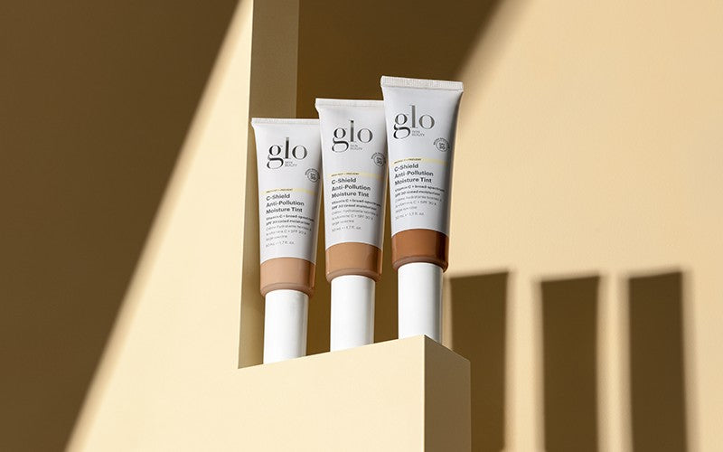 Three glo C-Shield Anti-Pollution Moisture Tint tubes in different shades on a beige pedestal with long shadows.